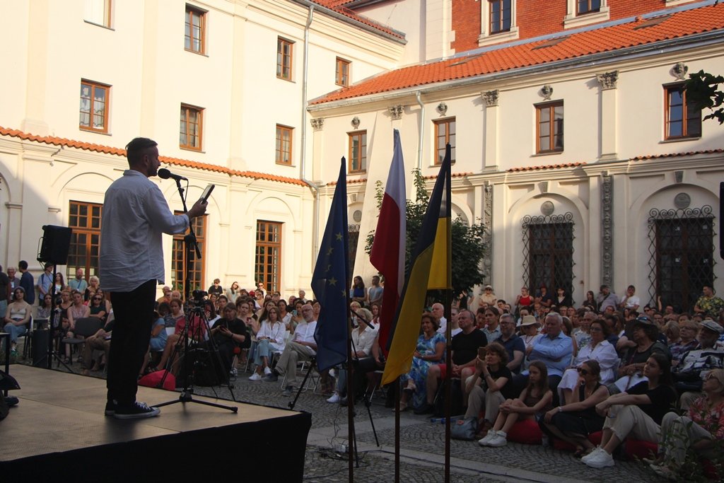 Na scenie mężczyzna mówi do siedzącej na dworze publiczności, po prawej stronie trzy flagi
